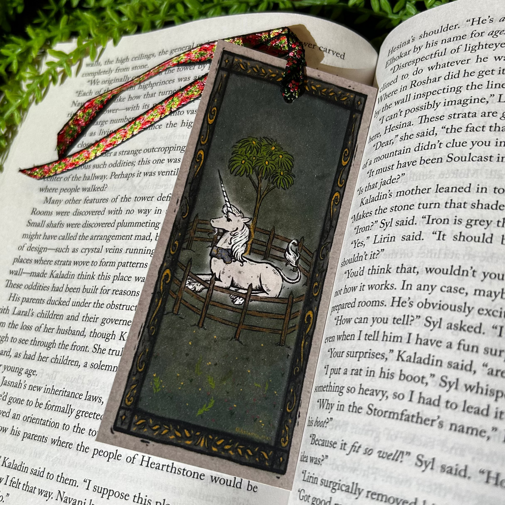 Decorative bookmark with unicorn design on an open book, surrounded by greenery and flowers.