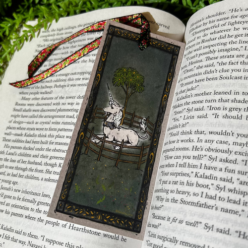 Decorative bookmark with unicorn design on an open book, surrounded by greenery and flowers.