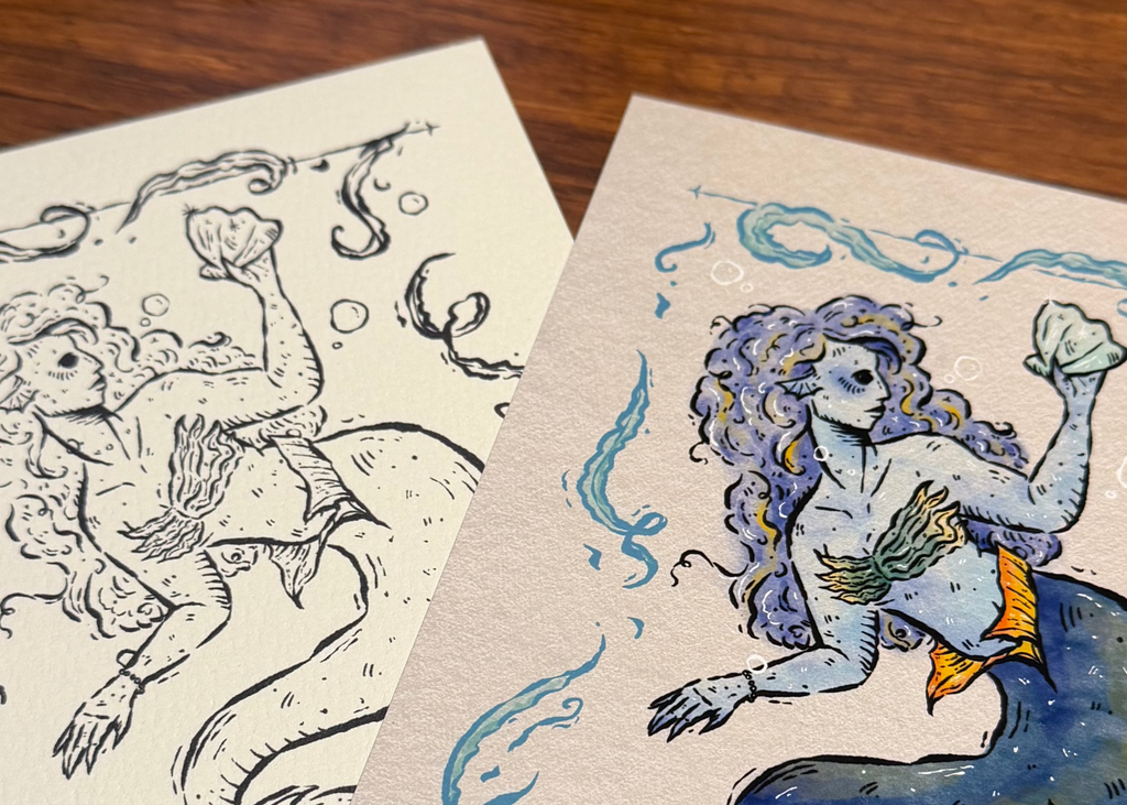 Two mermaid illustrations, one black and white and one colored, on a wooden surface with greenery.