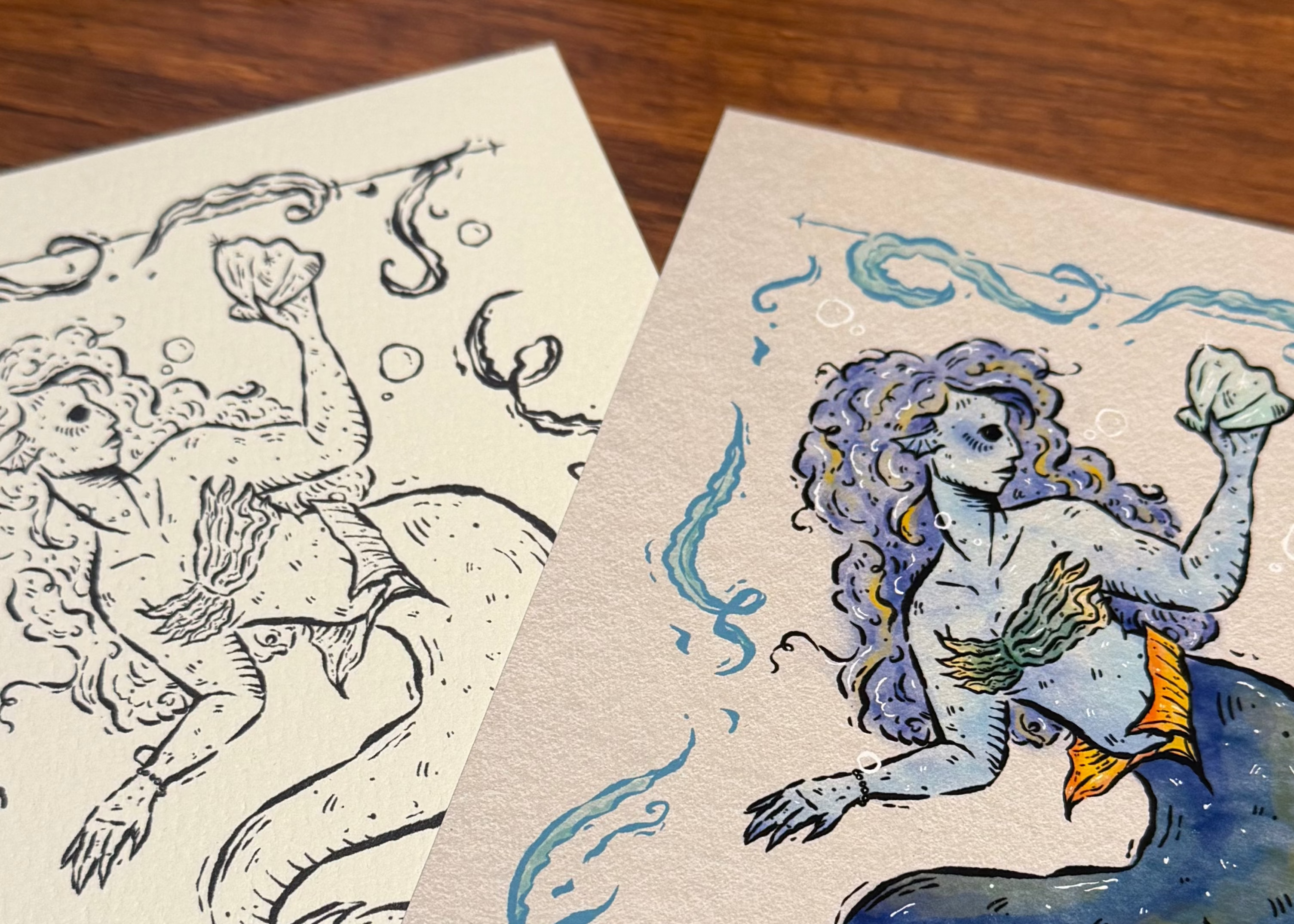 Two mermaid illustrations, one black and white and one colored, on a wooden surface with greenery.